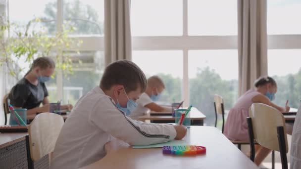 Student v ochranné masce studující ve škole během pandemie viru korony. Maskovaný učitel vysvětluje nové téma lekce maskovaným studentům. děti píšou perem ve třídě — Stock video