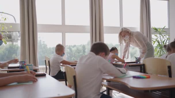 Studerande i skyddsmask som studerar i skolan under koronaviruspandemi. En maskerad lärare förklarar ett nytt lektionsämne för maskerade elever. barn skriver med pennor i klassrummet — Stockvideo