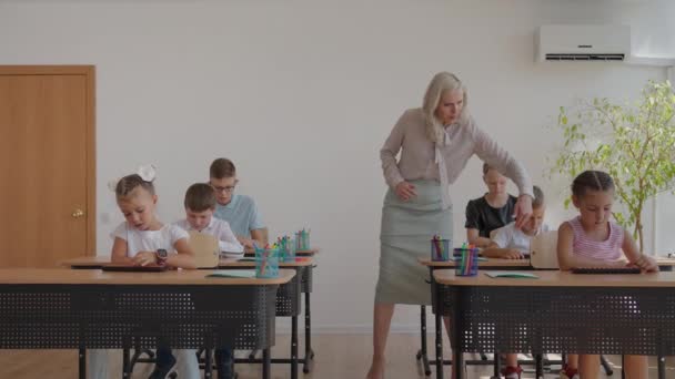 İlkokulda kadın öğretmen öğrenme sürecini kontrol ediyor. Bir bayan öğretmen masaların arasında yürür ve görevin tamamlanmasıyla ilgilenir. — Stok video