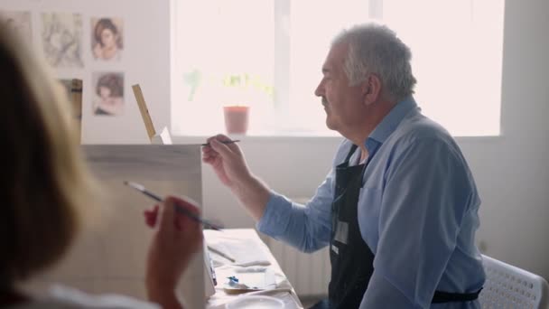 Una maestra le muestra a un hombre jubilado cómo dibujar un cuadro con pinturas y un pincel en los cursos para ancianos. Un hombre mayor dibuja un cuadro a un grupo de pensionistas — Vídeo de stock