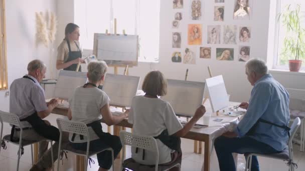 Öğretmen, yaşlılardan oluşan bir grup emekli insanın resim kurslarını gösteriyor. Bir grup yaşlı erkek ve kadın bir araya gelip gülümsüyor. — Stok video