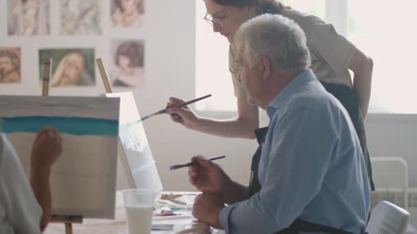 Μια γυναίκα δασκάλα δείχνει σε έναν συνταξιούχο πώς να ζωγραφίζει μια εικόνα με μπογιές και μια πινελιά στα μαθήματα για τους ηλικιωμένους. Ένας ηλικιωμένος ζωγραφίζει σε μια ομάδα συνταξιούχων. — Αρχείο Βίντεο
