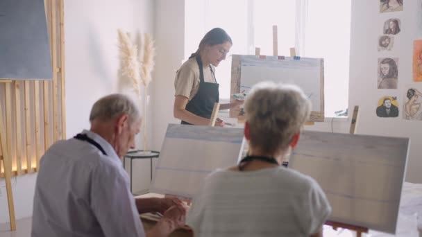 El profesor muestra a un grupo de amigos de jubilados de la tercera edad en cursos de dibujo. Un grupo de hombres y mujeres ancianos se reúnen y sonríen — Vídeo de stock