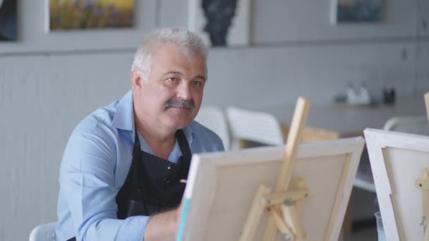 Uma professora mostra a um homem aposentado como desenhar um quadro com tintas e um pincel em cursos para idosos. Um idoso desenha um quadro para um grupo de pensionistas — Vídeo de Stock