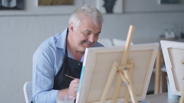 Uma professora mostra a um homem aposentado como desenhar um quadro com tintas e um pincel em cursos para idosos. Um idoso desenha um quadro para um grupo de pensionistas — Vídeo de Stock