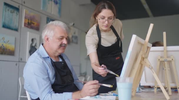 Hoge hoek uitzicht op vrolijke oudere vrienden schilderen op doek. Senior vrouw glimlachend terwijl ze tekent met de groep. Senioren wonen samen schilderles bij. Oudere mannen die plezier hebben met schilderen in de kunstklas — Stockvideo