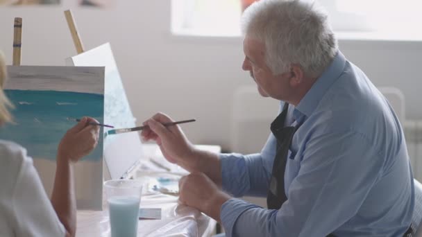 A female teacher shows a retired man how to draw a picture with paints and a brush at courses for the elderly. A senior man draws a picture to a group of pensioners — Stock Video