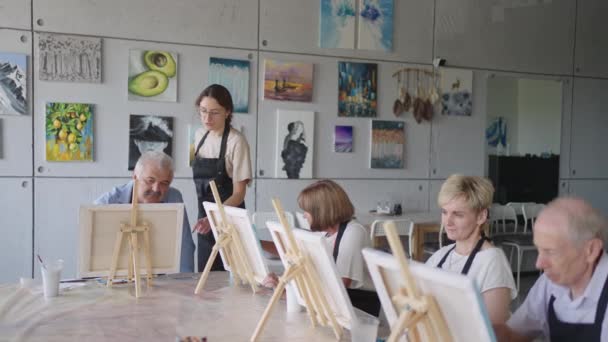 Вид сбоку счастливых пожилых людей, улыбающихся во время рисования в качестве рекреационной деятельности или терапии в классе рисования вместе с группой пенсионеров и мужчин — стоковое видео