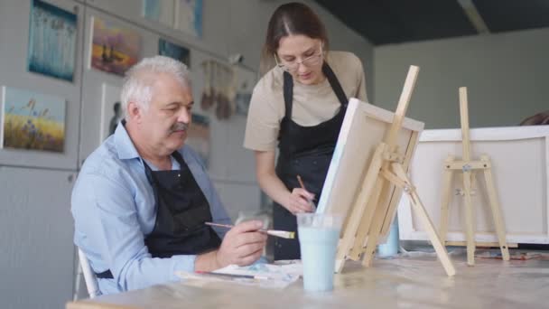 En kvinnlig lärare visar en pensionerad man hur man ritar en bild med färger och en pensel på kurser för äldre. En äldre man ritar en bild till en grupp pensionärer — Stockvideo