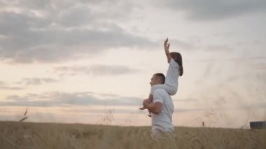 Babası, sevgili sağlıklı kızını güneşte omuzlarında taşıyor. Yavaş çekimde, kız babasıyla sahada yürüyor ve özgür ve mutlu bir şekilde ellerini kaldırıyor. Sahada yürümek.