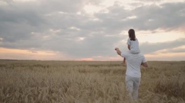 Babacığın sevgili, sağlıklı kızını güneşte omuzlarında taşıyor. Yavaş çekimde, kız babasıyla birlikte sahada yürüyor ve özgür ve mutlu bir şekilde ellerini sallıyor.. 