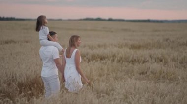 Yavaş çekim: Çocuklu mutlu çiftçi ailesi buğday tarlasında yürüyor. Sağlıklı anne, baba ve küçük kız birlikte doğanın tadını çıkarıyorlar..