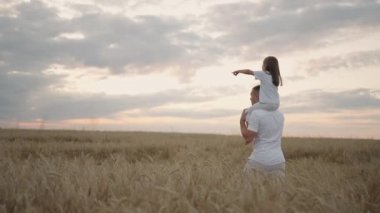 Babası, sevgili sağlıklı kızını güneşte omuzlarında taşıyor. Yavaş çekimde, kız babasıyla sahada yürüyor ve özgür ve mutlu bir şekilde ellerini kaldırıyor. Sahada yürümek.