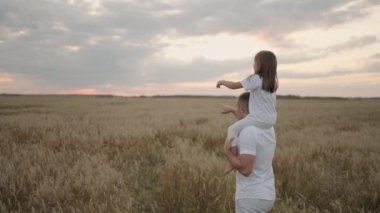 Babacığın sevgili, sağlıklı kızını güneşte omuzlarında taşıyor. Yavaş çekimde, kız babasıyla birlikte sahada yürüyor ve özgür ve mutlu bir şekilde ellerini sallıyor.. 