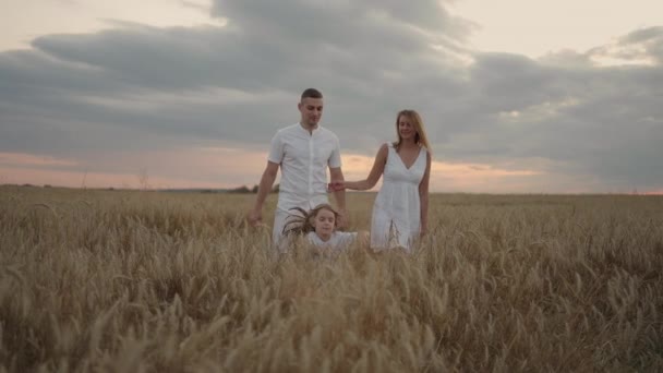 Young couple of parents with girl children holding hands of each other and running through wheat field at sunset. Happy family jogging among barley meadow and enjoying nature together. Slow motion — Stock Video