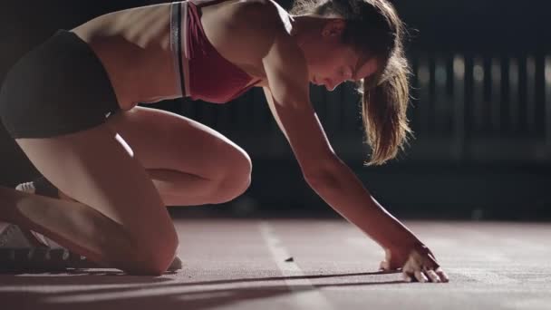 Kvinnliga idrottare utbildning på löparbanan i den mörka stadion. Sakta i backarna. En ung kvinnlig idrottsman kliver in i dynorna och börjar i loppet. närbild av en flicka löpare — Stockvideo