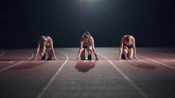 En rad löpare kvinnor huka i utgångsläget innan de börjar tävla. Kvinnor börjar med löparskor på arenan från startlinjen i mörker med spotlights i slow motion. — Stockvideo