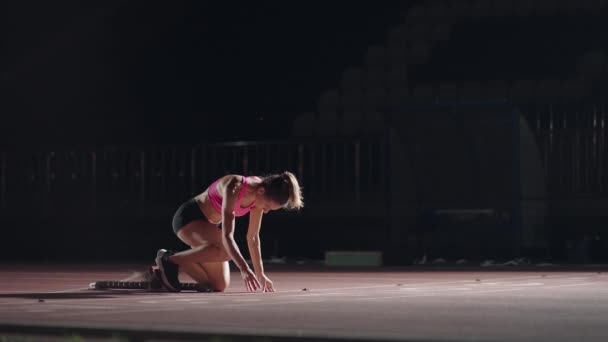 Açık hava spor stadyumundaki bir koşu pistindeki karışık ırk bayan sporcusu başlangıç bloklarından başlayarak, yavaş çekimde lens aleviyle arka planda bir yarış başlatıyor. Gece — Stok video