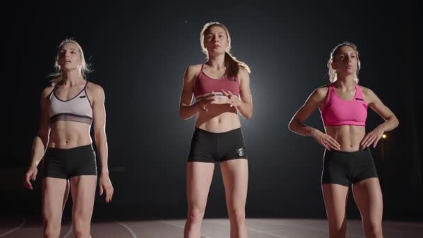 Three women athletes prepare for a track race in a dark stadium with streetlights on. Time-lapse footage of warm-up and concentration of a group of women before the race on the track — Vídeo de stock