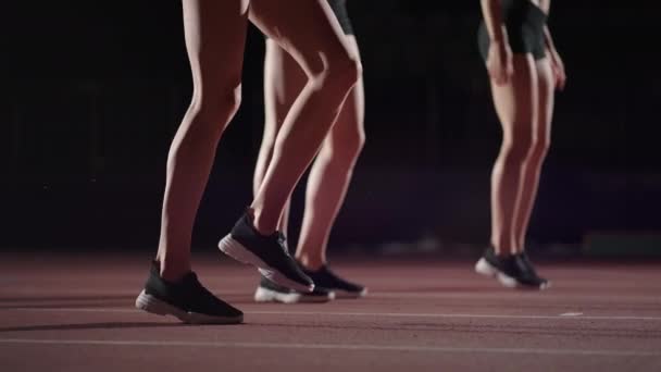 Три спортсменки готовятся к бегу на тёмном стадионе с включёнными уличными фонарями. Замедленная съемка разминки и концентрации группы женщин перед гонкой на трассе — стоковое видео