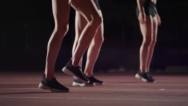 Three women athletes prepare for a track race in a dark stadium with streetlights on. Time-lapse footage of warm-up and concentration of a group of women before the race on the track — Stock Video