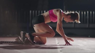 Açık hava spor stadyumundaki bir koşu pistindeki karışık ırk bayan sporcusu başlangıç bloklarından başlayarak, yavaş çekimde lens aleviyle arka planda bir yarış başlatıyor. Gece