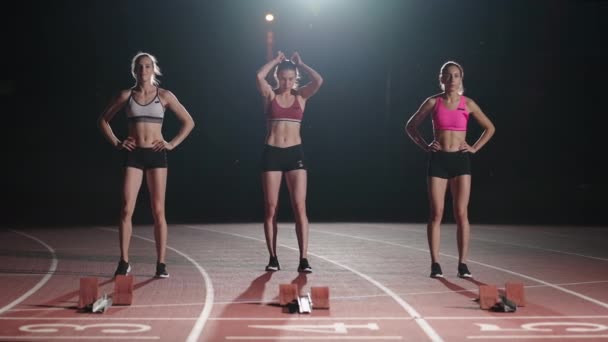 Three women athletes prepare for a track race in a dark stadium with streetlights on. Time-lapse footage of warm-up and concentration of a group of women before the race on the track — Stockvideo