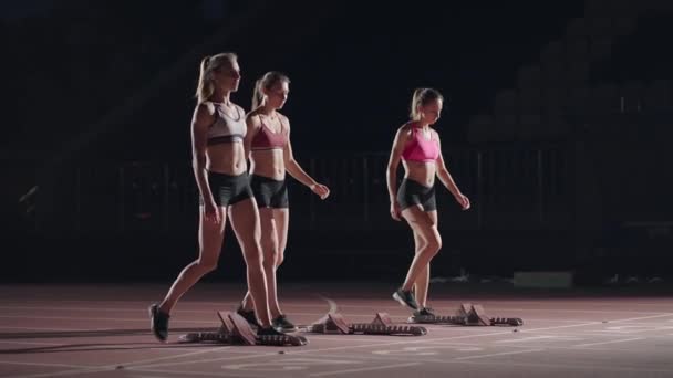 A row of runners womens crouch in the starting position before beginning to race. Females start with running shoes on the stadium from the start line in the dark with spotlights in slow motion. — Stockvideo