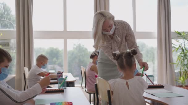 I skolan, i klassrummet, skriver barn anteckningsböcker i skyddsmasker, observerar ett socialt avstånd. Skolutbildning under pandemin — Stockvideo