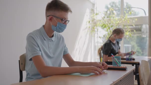 À l'école, en classe, les enfants écrivent dans des cahiers portant des masques protecteurs, observant une distance sociale. Éducation scolaire pendant la pandémie — Video