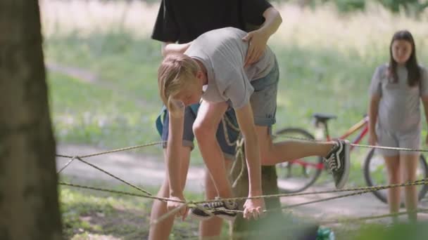 在一位教孩子攀岩和旅游的导游的帮助下，夏令营徒步旅行的孩子们正沿着绳索走着。森林里的一个男孩冲破了绳索屏障 — 图库视频影像