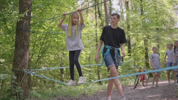 Klein meisje in klimuitrusting in een touwenpark. Een groep Kaukasische kinderen trainend op het trainingskamp. In het kinderkamp worden kinderen geleerd obstakels te overwinnen met behulp van een touwoversteek. — Stockvideo