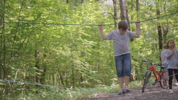 Liten pojke i klätterutrustning i en rep Park. Grupp kaukasiska barn utbildning på boot camp. I barnens läger får barnen lära sig att övervinna hinder med hjälp av en repkorsning. — Stockvideo