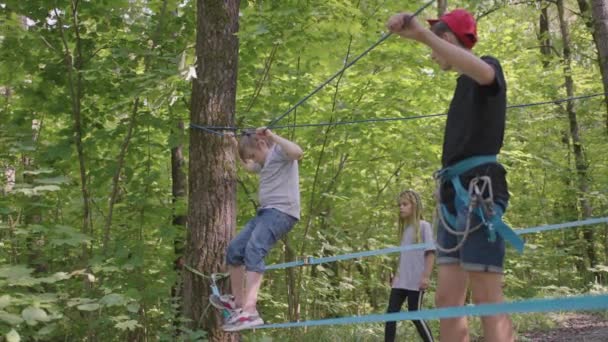 Kinderen op een zomerkamp wandelen langs de touwen met behulp van een gids die kinderen rotsklimmen en toerisme leert. Een jongen in het bos overwint een touw barrière — Stockvideo