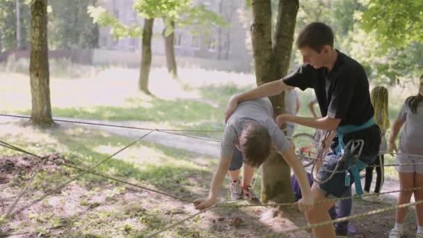 Kinderen op een zomerkamp wandelen langs de touwen met behulp van een gids die kinderen rotsklimmen en toerisme leert. Een jongen in het bos overwint een touw barrière — Stockvideo