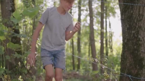 Um menino em uma caminhada de acampamento de verão supera um obstáculo rastejando no chão e caindo. Divertidos desafios das crianças — Vídeo de Stock