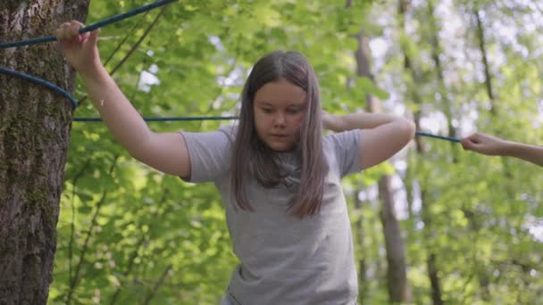 Bambino attivo scala senza paura le corde tra gli alberi. Ragazza in un parco avventura sono passare ostacoli sulla strada corda. campeggio per bambini, campo estivo, — Video Stock