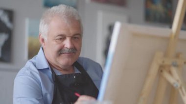 Önlüklü yaşlı bir adam masada otururken fırçayla bir resim çizer.