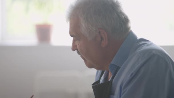 Een oudere man in een schort schildert een schilderij met een borstel terwijl hij aan tafel zit — Stockvideo