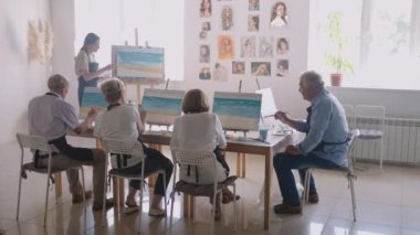 Bir grup yaşlı erkek ve kadın arkadaş birlikte izleyip resim tekniğini öğretmenin arkasında tekrarlıyorlar.