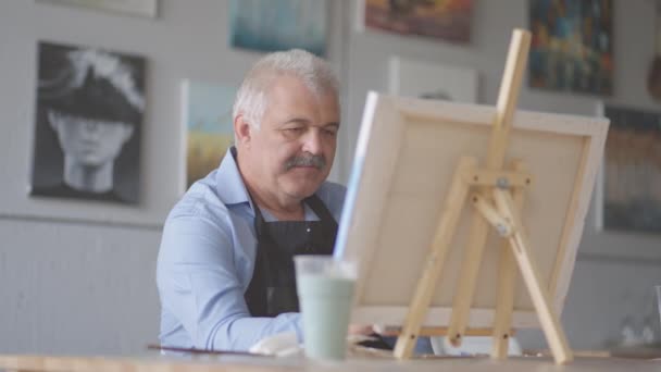 Egy idős férfi kötényben ecsettel fest egy képet, miközben az asztalnál ül. — Stock videók