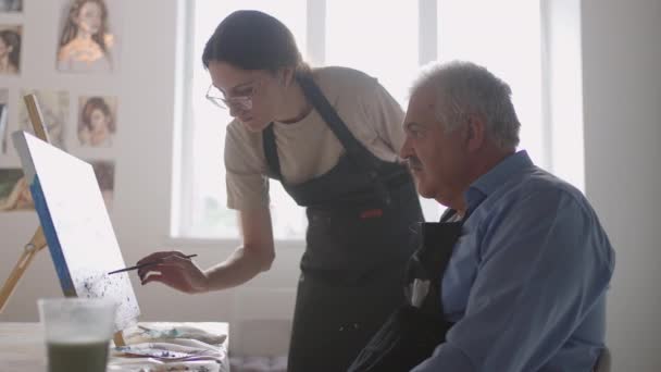 Genç bir kadın öğretmen yaşlı bir emeklinin resim yapmasını öğrenmesine yardım ediyor. Kıdemli adam resim yapmayı öğreniyor. — Stok video