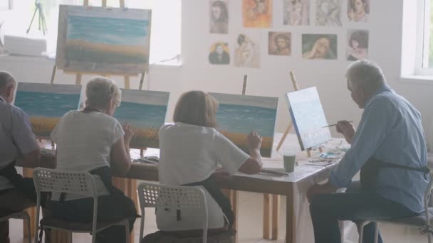 Uma jovem professora de arte demonstra a técnica de pintura com tintas acrílicas sobre tela para um grupo de aposentadas. Grupo de homens e mulheres seniores — Vídeo de Stock