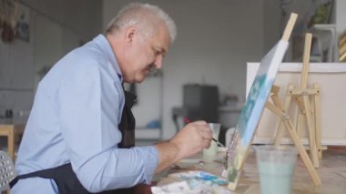 Önlüklü yaşlı bir adam masada otururken fırçayla bir resim çizer.