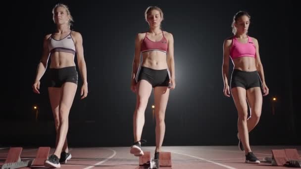 Jóvenes mujeres bonitas corriendo en ropa deportiva y zapatillas de deporte preparándose para la carrera en la pista de jogging juntos. Comienza desde la línea y comienza la carrera en cámara lenta en la oscuridad — Vídeos de Stock