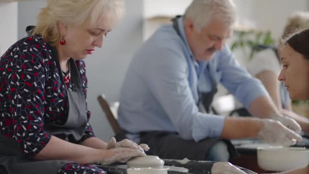 Medium shot van middelbare leeftijd keramische kunstenaar onderwijs groep ouderen Kaukasische vrouw en senior man hoe om klei wig zitten aan het bureau in kunststudio. Praten op het werk — Stockvideo