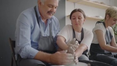 Orta yaşlı seramik ressamın yaşlı beyaz kadınlara ve üst düzey adama sanat stüdyosundaki masada otururken kilin nasıl sıkıştırılacağını öğretirken orta yaşlı bir fotoğrafı. İnsanlar iş yerinde konuşmaktan hoşlanıyor.