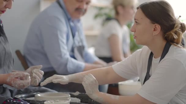 Een vrouwelijke pottenbakkers wiel beeldhouwen leraar legt uit hoe te werken en leert een oudere vrouw om te werken met klei en maken mokken en kannen. Masterclass voor gepensioneerden. Aardewerk cursussen — Stockvideo