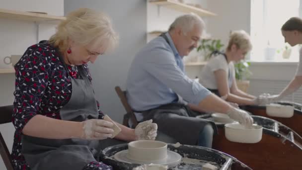 Een vrouwelijke pottenbakkers wiel beeldhouwen leraar legt uit hoe te werken en leert een oudere vrouw om te werken met klei en maken mokken en kannen. Masterclass voor gepensioneerden. Aardewerk cursussen — Stockvideo