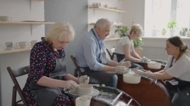 Orta yaşlı seramik ressamın yaşlı beyaz kadınlara ve üst düzey adama sanat stüdyosundaki masada otururken kilin nasıl sıkıştırılacağını öğretirken orta yaşlı bir fotoğrafı. İnsanlar iş yerinde konuşmaktan hoşlanıyor.